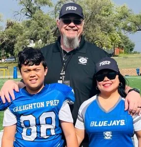 Thom Sisson with his family on Football Sunday — Thom in his coach’s uniform, his son in full pads, and his wife cheering in team gear — celebrating faith, family, and teamwork.
